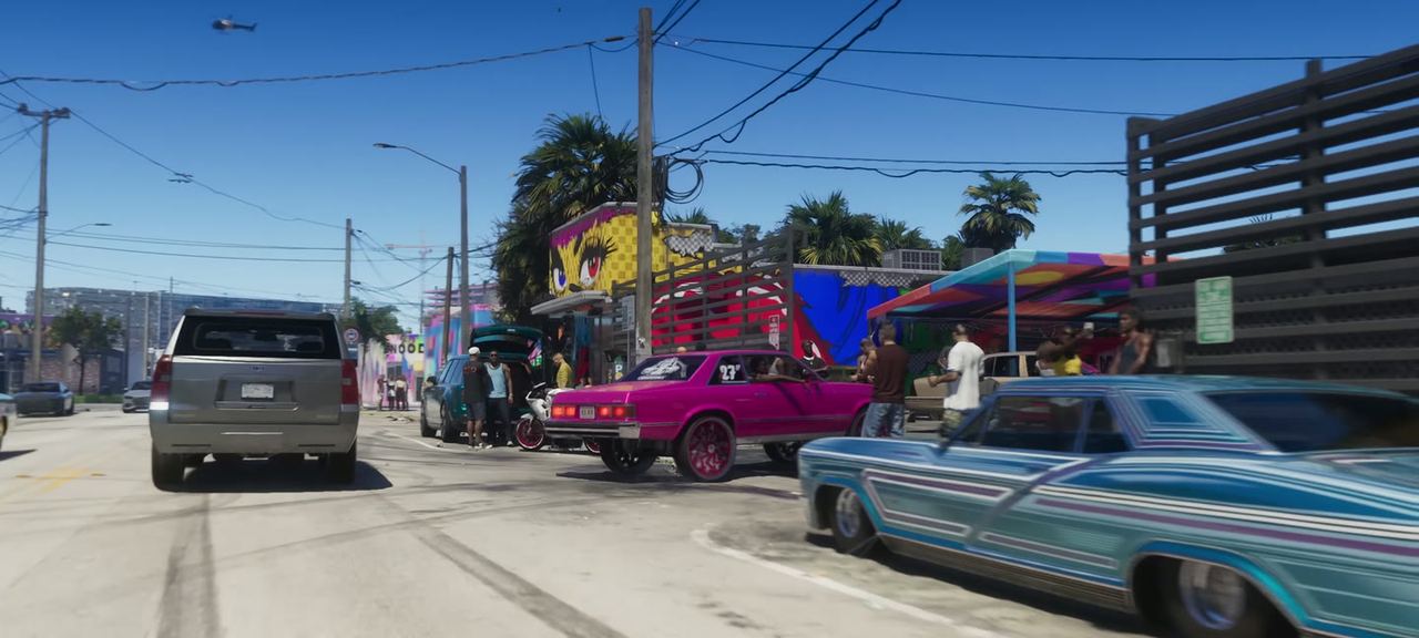 A street of shopfronts with a blue classic sedan on whitewalls, a pink coupe and a van.